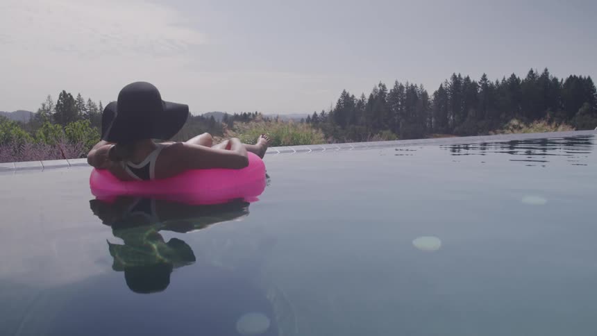 A woman relaxes with a drink while floating on an inflatable ring in an infinity pool