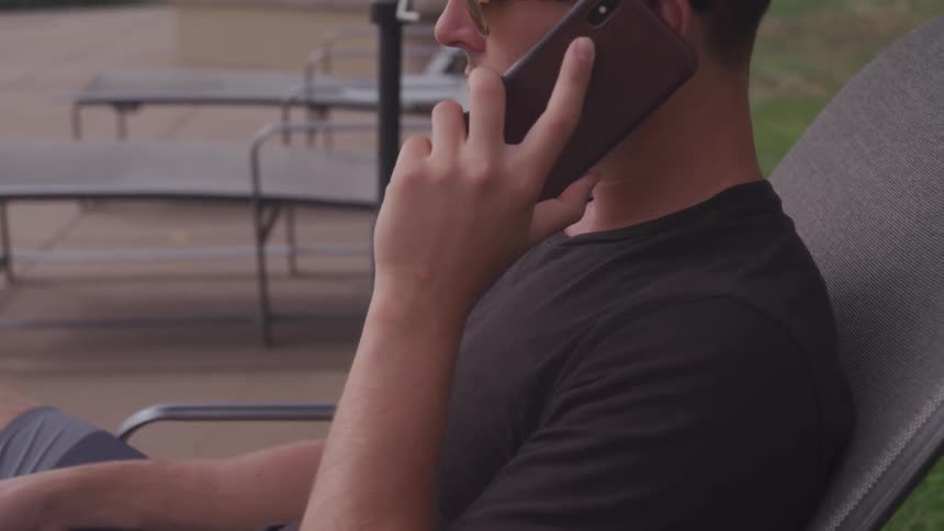 A white man sits on a deck chair and talks on a smartphone