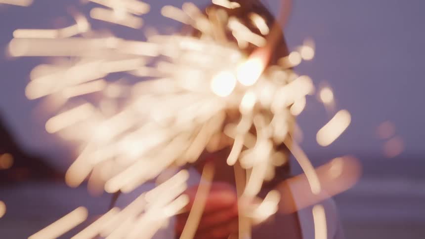 A woman wearing a loose white top holds a sparkler on the beach at night
