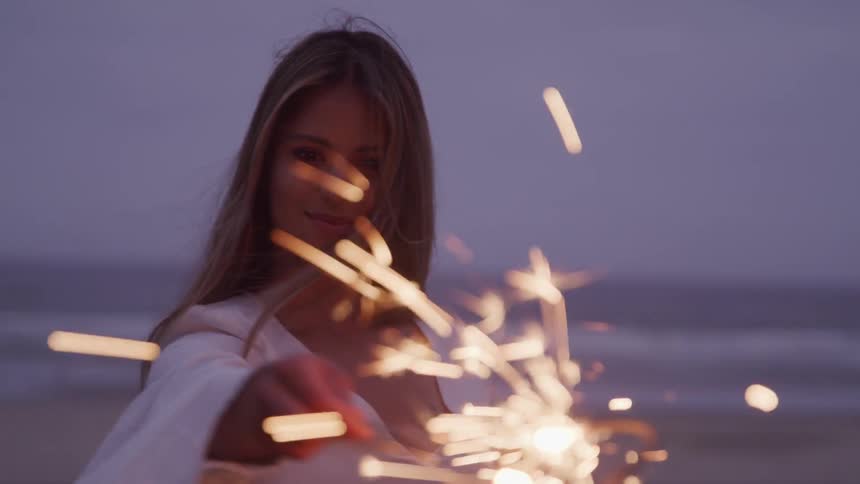 A woman wearing a loose white top holds a sparkler on the beach at night