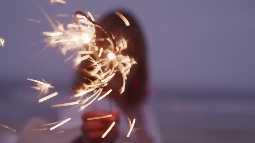 A woman wearing a loose white top holds a sparkler on the beach at night