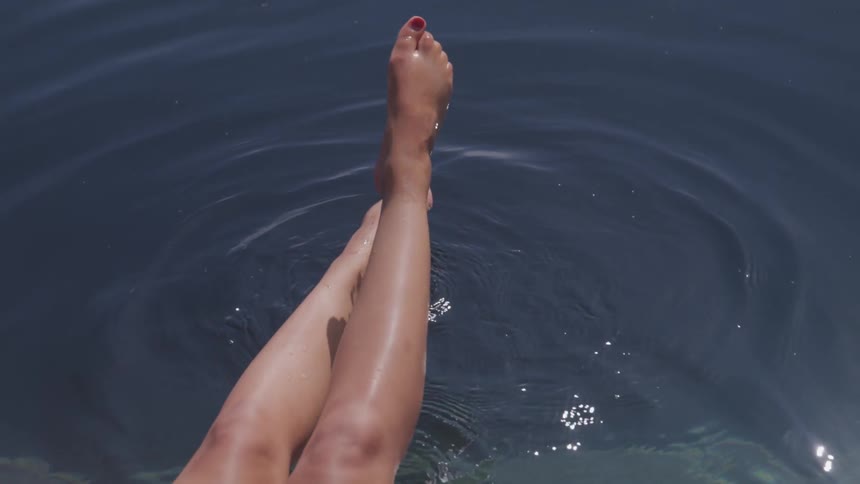 A woman sits on the edge of a pool waving and splashing her legs and feet