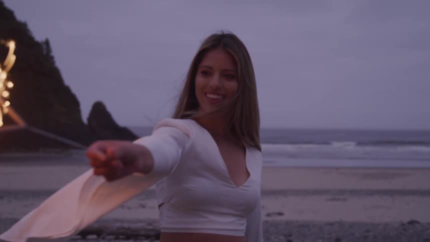 A woman wearing a loose white top holds a sparkler on the beach at night