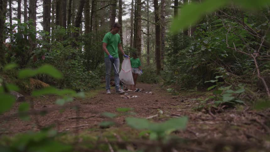 A man and woman wearing green shirts are picking up trash in the forest along a dirt trail