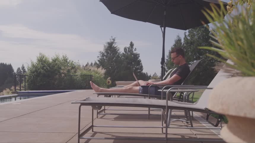A man sits beside a pool and types on his laptop
