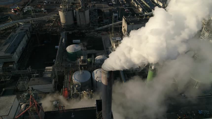 At a large industrial facility a towering smoke stack releases a thick cloud of smoke into the air