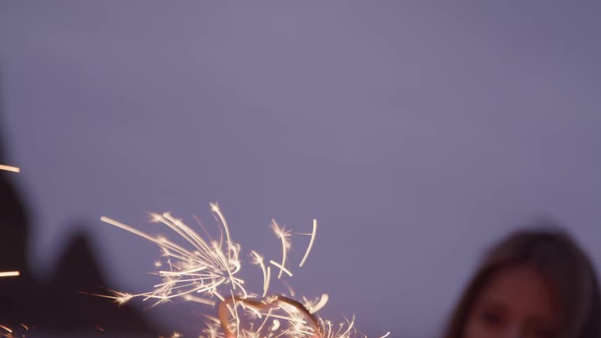 A woman wearing a loose white top holds a sparkler on the beach after sunset