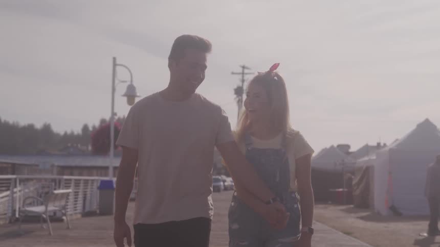 A young couple smile as they holds hands and walk along a concrete walkway on a sunny day