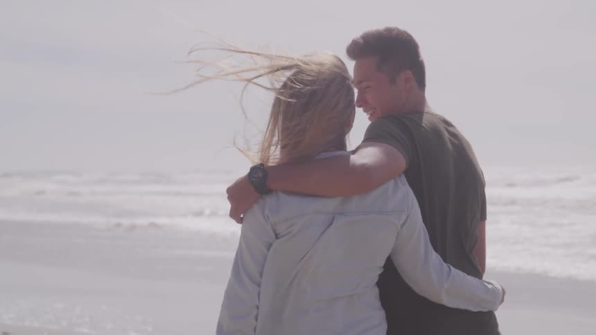 A man and woman walk along the beach with their arms around one another