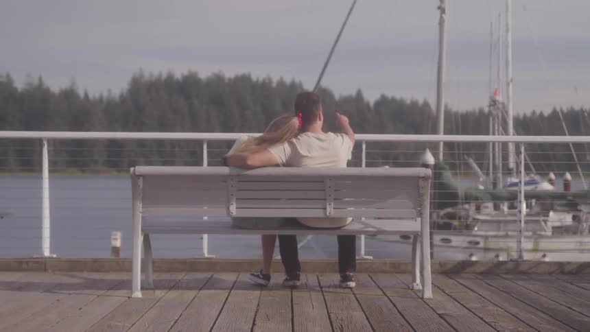Man and woman sit on a bench on a pier and look out at the sailboats in the water