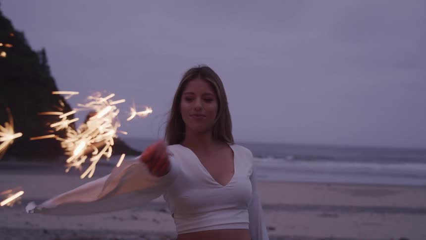 A woman wearing a loose white top holds a sparkler on the beach at sunset