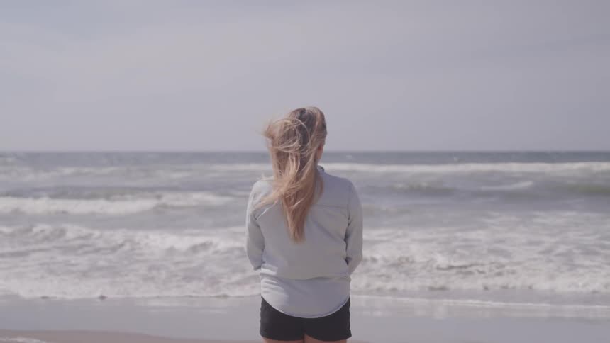 A woman in short shorts staring out to sea on a windy day