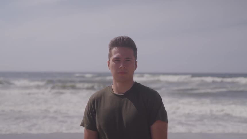 A man stands on a beach with his back to the ocean as waves come to shore