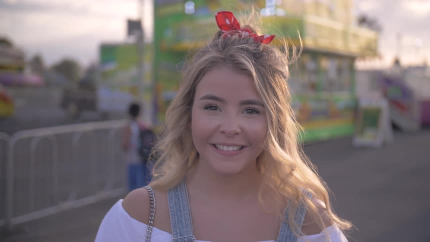 A young woman smiles while standing outside at an fairground where there are rides behind her