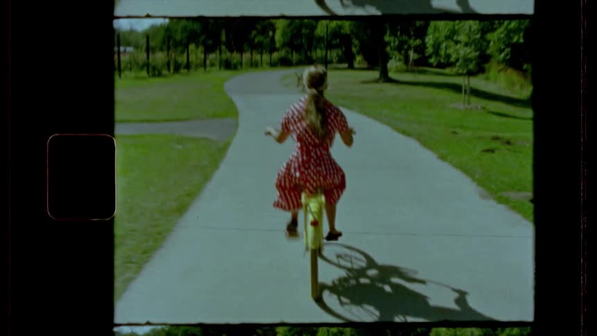 Female wearing a red dress with white polka dots rides a yellow bicycle along a paved pathway through the park