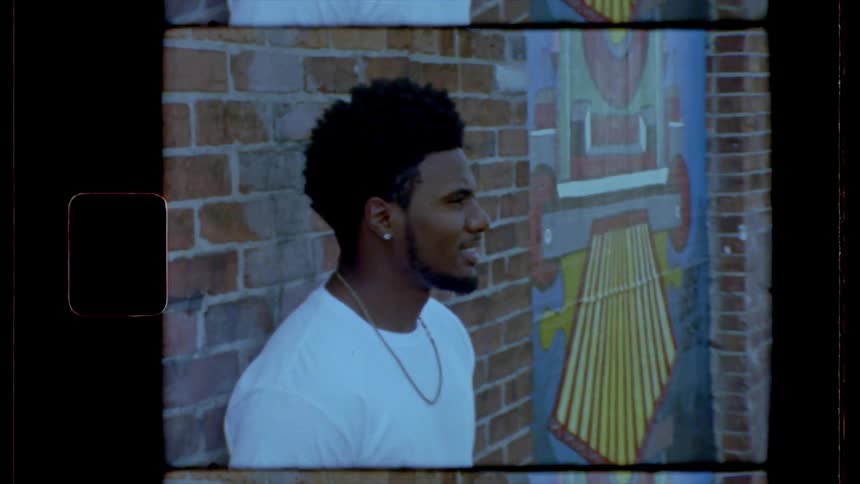 Black man wearing a white t shirt leans against the brick wall of the building