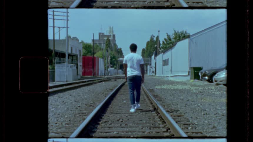 A man wearing a white tee shirt and jeans walks down railroad tracks