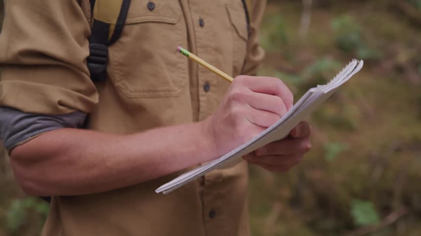 A mans forearm as he stands and makes notes using a pencil while outdoors in nature