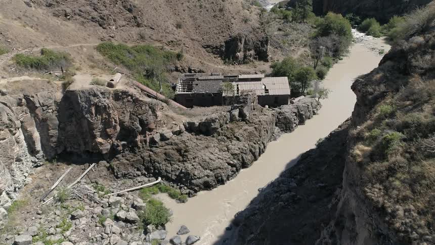 A broken down building sits above a muddy river in a canyon