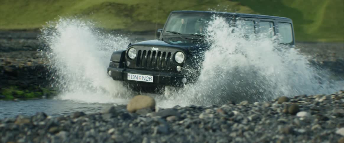 A jeep splashes through a stream at the base of a grassy slope