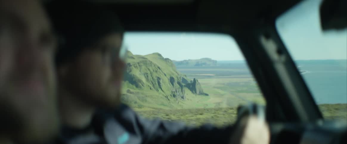 Two white males are driving in a mountainous area with an ocean in the background