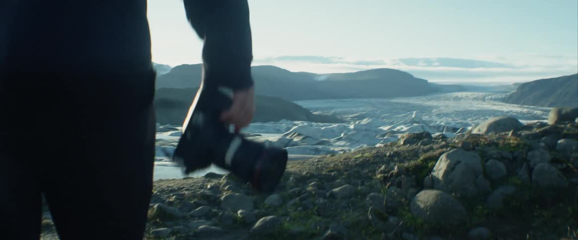 A photographer carries a camera as he walks across a grassy area that overlooks the coast