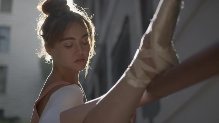 A young female ballerina has her foot placed atop a railing to and stretches while standing outdoors surrounded by tall buildings