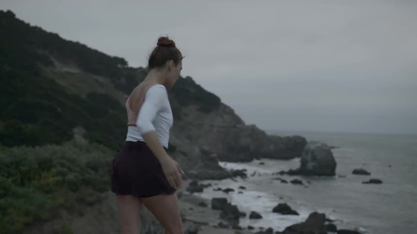 Ballerina reaches up the cloudy sky with one leg forward and the other leg back on the hill by the seaside on a windy day