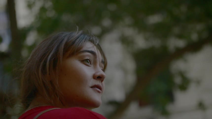A young woman standing outdoors near a building and a tree looks out at her surroundings