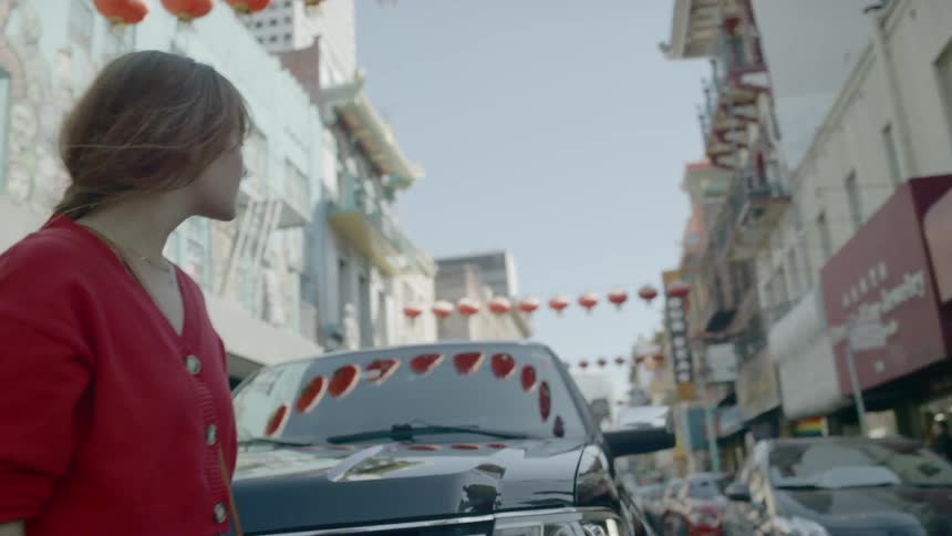 A woman in a red shirt runs her hand through her hair as she crosses in front of a stopped car in the road
