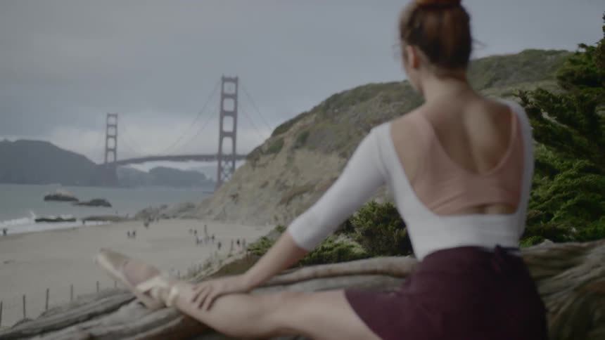 A young ballerina has her foot placed atop a fallen tree while stretching her leg outdoors with the golden gate bridge in the background over the ocean water