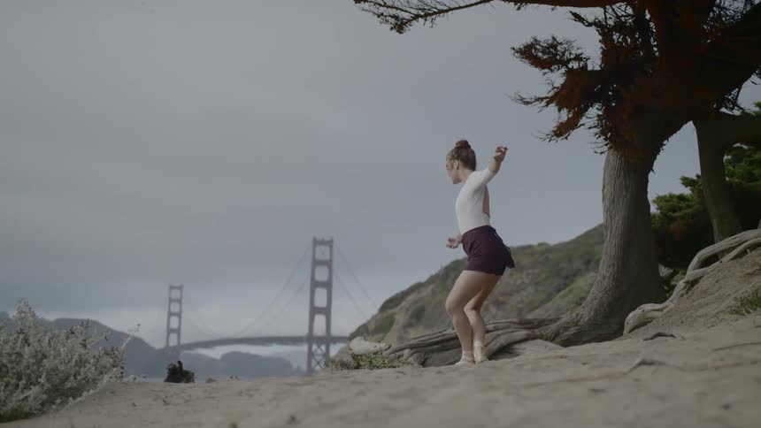 Woman performing ballet pose on a cliff with bridge in the distance