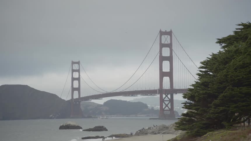 People walk on a beach while the golden gate bridge sits in the background on a typical foggy day