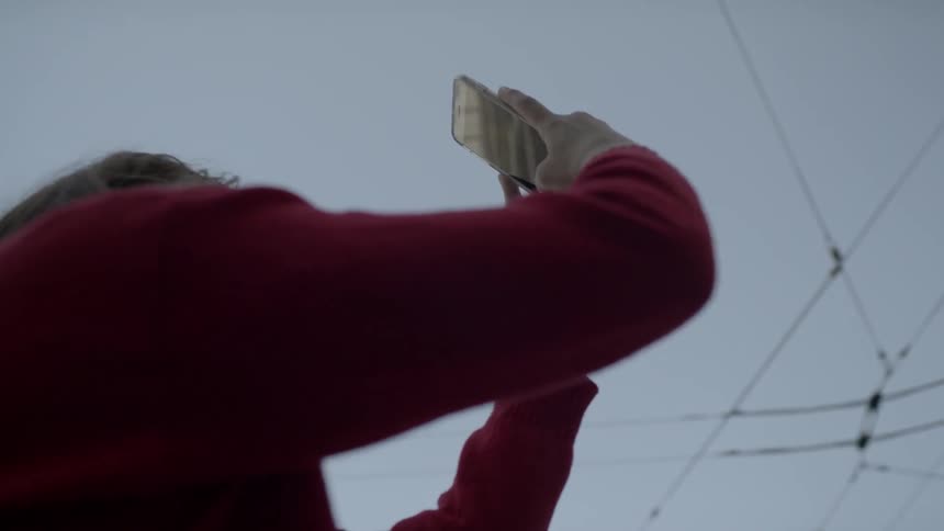 A woman standing at a city intersection holds her phone up high to photograph a building