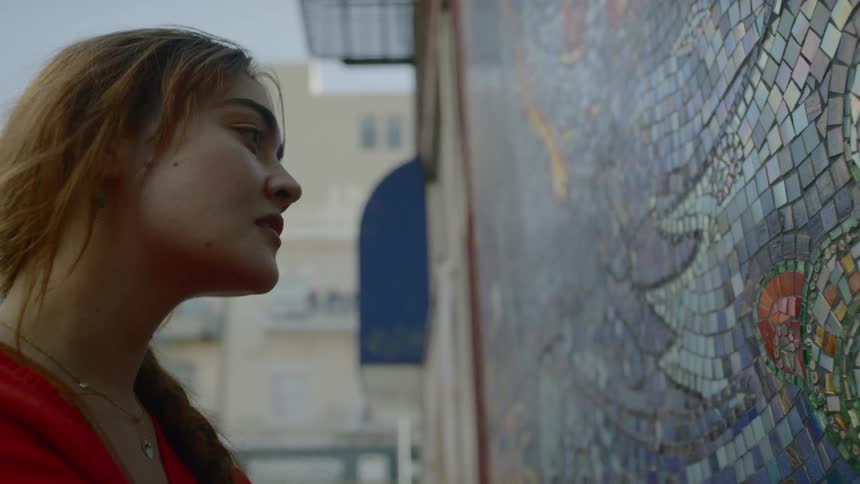 A young woman stands next to a colorful mosaic mural made with countless small tiles