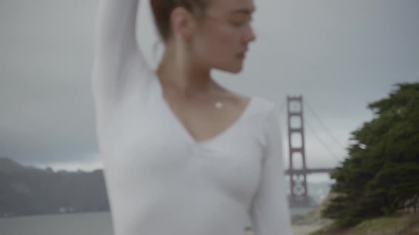 A young ballerina performs ballet dance moves outdoors near the coast with the golden gate bridge behind her in the distance