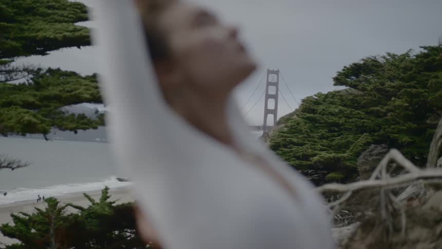 A woman stretches on a beach surrounded by trees with the golden gate bridge in the background
