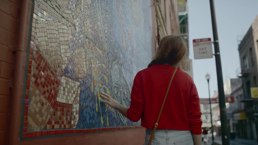 A woman walking down a city street stops in front of a tiled mural on a building