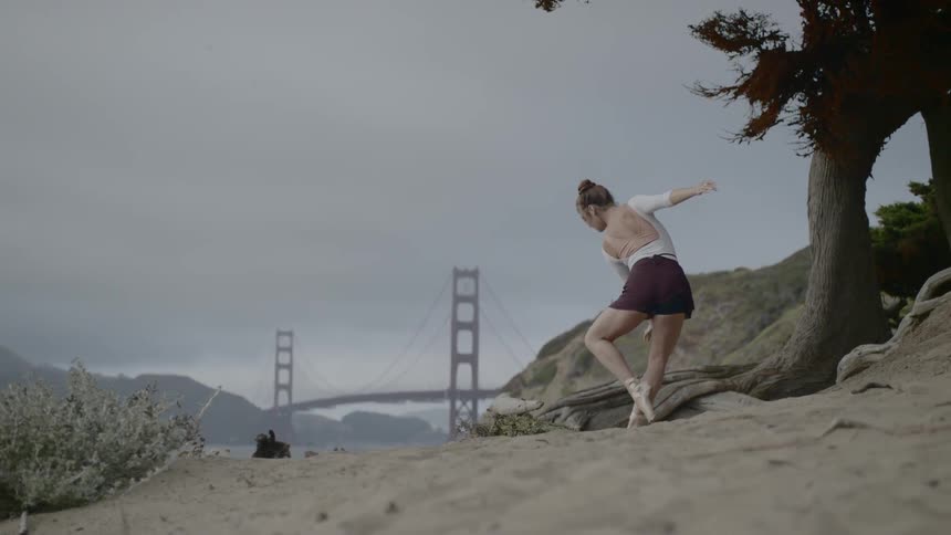 Balerina wearing toe shoes dances outdoors on beach with suspension bridge in background