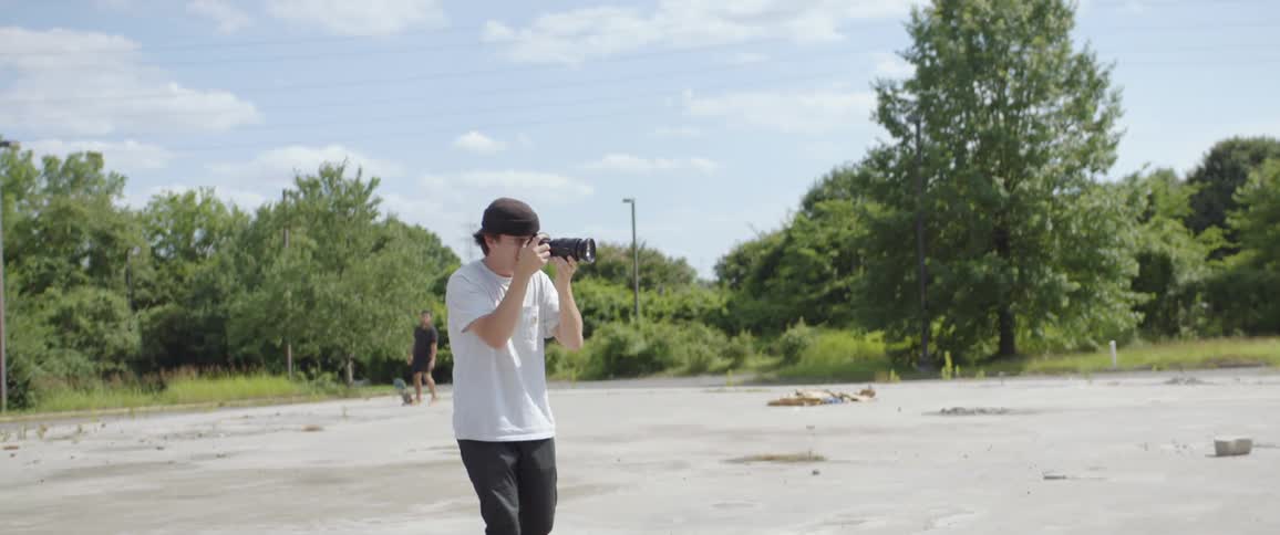 A man looks through a camera with a large lens as he walks backwards on a concrete parking lot