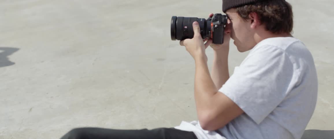 Male photographer wearing a brown beanie hat lies on the ground and takes picture of people riding a skateboard outdoors