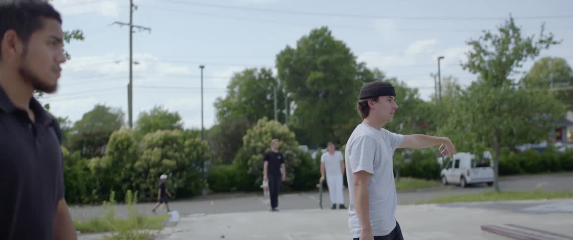 A man in a hat points forwards while his friends skateboard on a sunny day