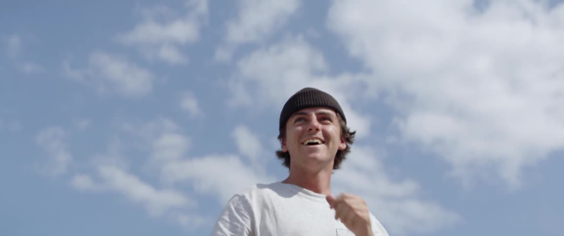 Male photographer wearing a brown beanie hat laughs as he gets up off the ground to take picture of people riding a skateboard outdoors
