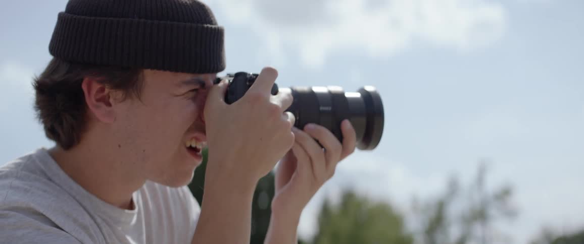 A young white man follows a subject with their camera on a bright day under a blue sky filled with puffy clouds