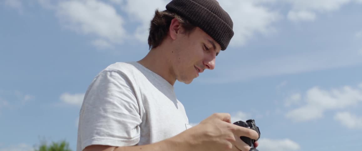 A teenager checks photos on his digital camera as people skateboard behind him
