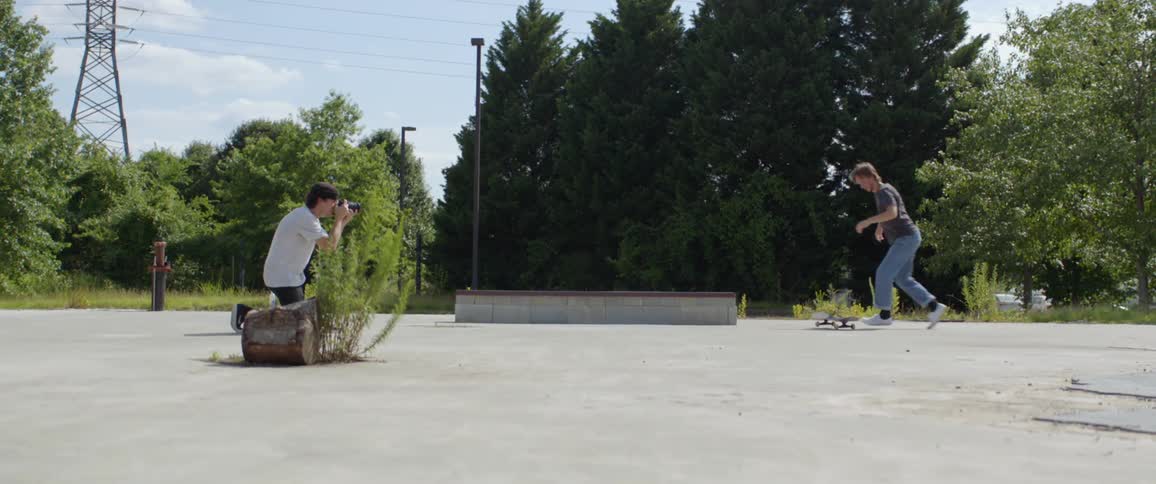 A man kneels on the pavement to photograph a skateboarder doing a trick at a skate park