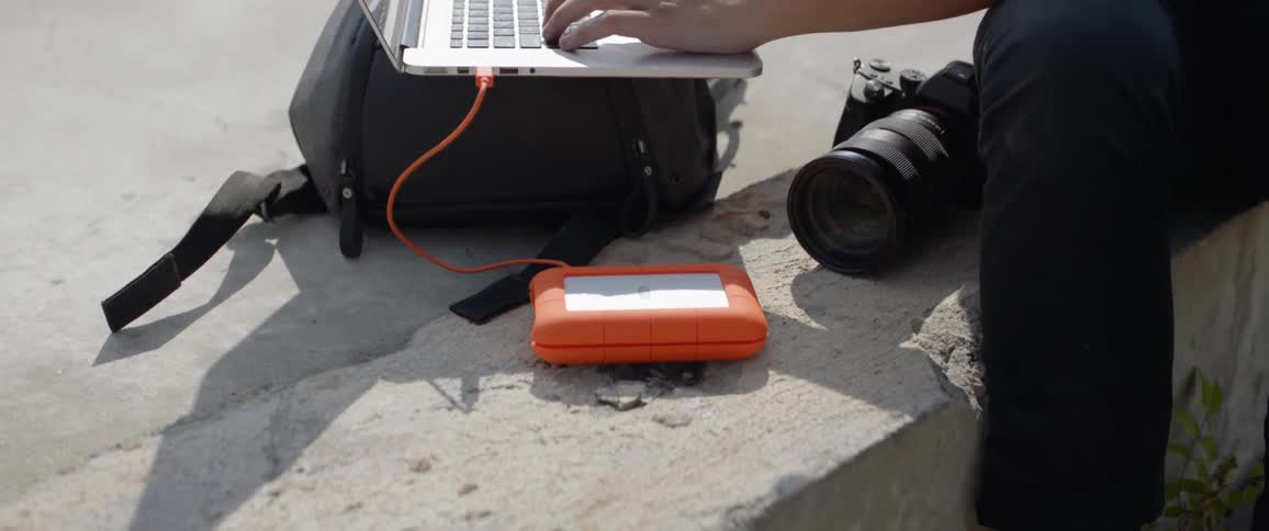 A person sits on a wall typing on a laptop resting on a camera bag with camera and lens beside it