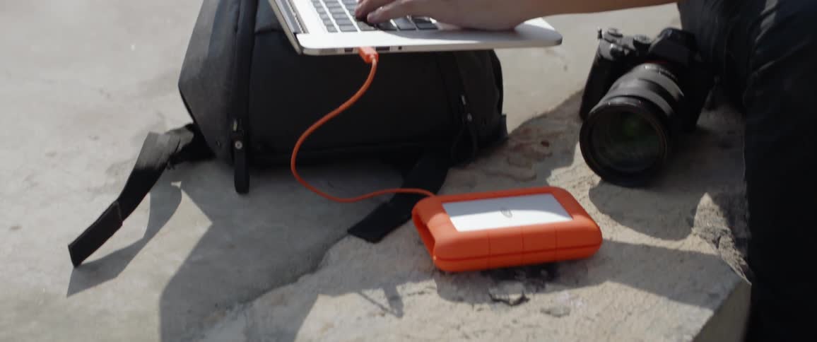 A man sitting on concrete typing on his laptop thats on a bag