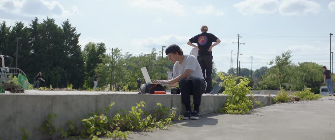 A teen sits outside and types while others talk and ride skateboards