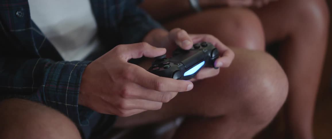 A man sitting on a couch uses a wireless controller to play a video game